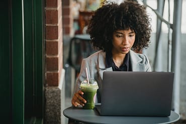 attentive black businesswoman watching laptop with beverage in cafe